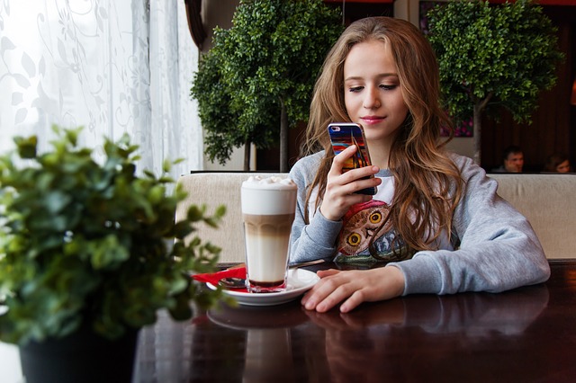 A teenage girl sitting in a restaurant, smiling as she texts something on her phone.